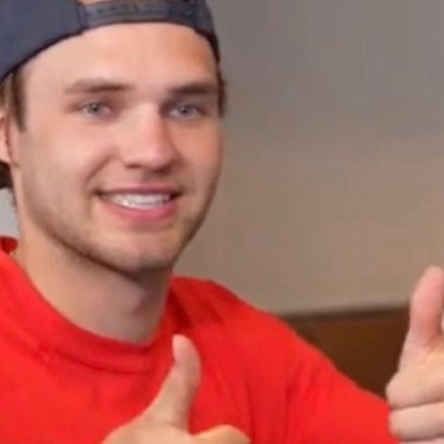 Close-up picture of Josh Norris as he holds two thumbs up, while he smiles with his teeth clenched. He’s wearing a red T-shirt and a backwards black baseball cap. 