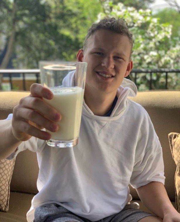 Brady Tkachuk sits on a sofa on a porch outside. He’s wearing a white short-sleeved hoodie and grey shorts. He’s holding a glass of milk up towards the viewer. 