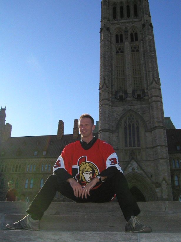 Dominik Hašek sits on the steps in front of the Parliament Building in Ottawa, with his legs bent and spread apart. He’s wearing a red Sens jersey with the Senagoth logo over a black stripe; the red extends past the shoulders, and towards white and then black parts on the arms. The number 39 is in red on the white sections. He’s also wearing black pants and grey-ish laced shoes. The sky in the background is clear and light blue. 