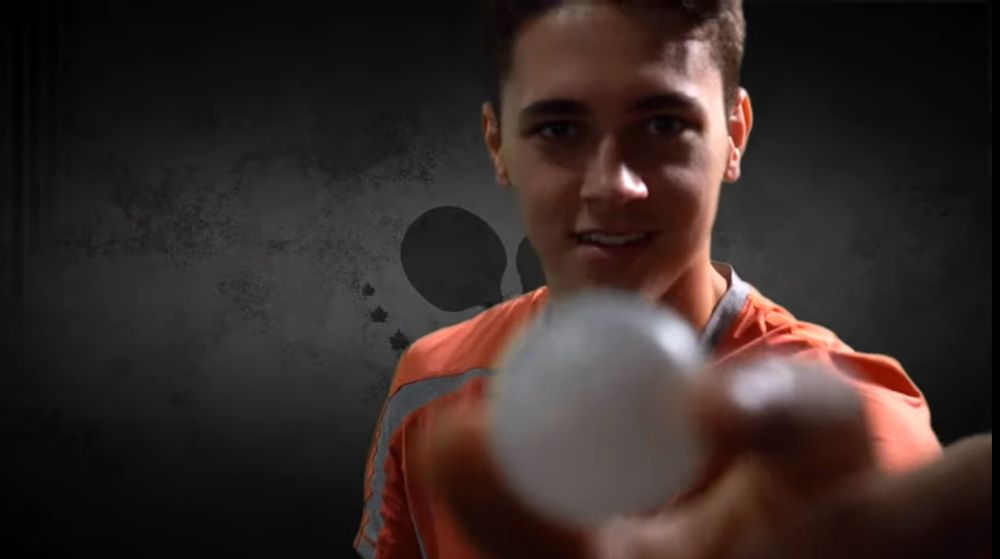 Shane Pinto stands in front of a grey background, holding out a white golf or ping pong ball to the viewer. He’s wearing an orange shirt with a grey stripe travelling from the right side of his neck across his left shoulder. The ball is unfocused in comparison to him. 