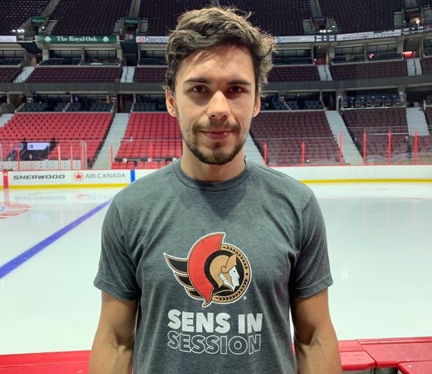 Artem Zub stands by the bench at the CTC, with the ice rink and stands visible behind him. He’s wearing a grey T-shirt with a Sens logo, and the text “Sens In Session” in all-caps beneath it. The first two words are solid white, and the last one is outlined in white. He’s visible from the waist up, with his arms by his side. He has short wavy brown hair and a mustache and facial hair on his chin. 
