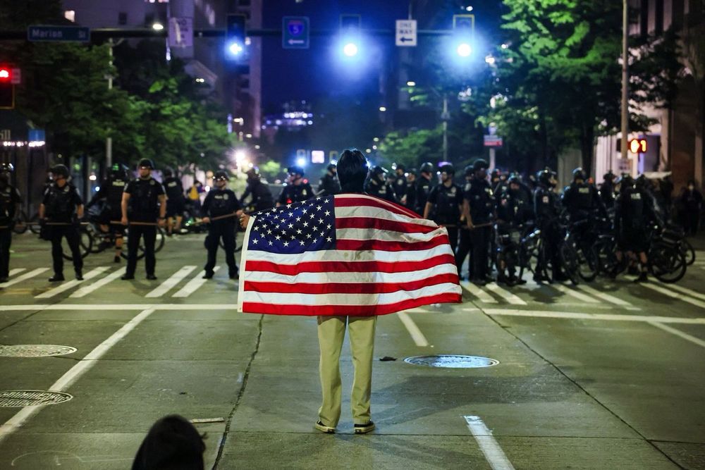 Person draped in an American flag stands before a line of riot police