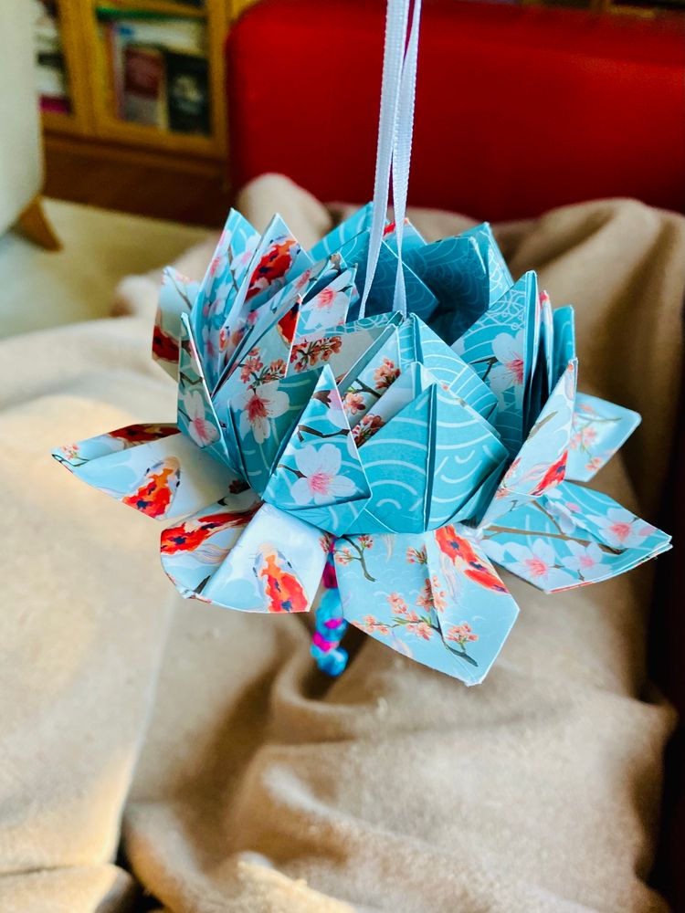 Schräger Blick von oben: Eine aus Papier gefaltete Lotus-Blume mit Band zum Aufhängen. Das Papier ist zart hellblau/rosé mit Kirschblüten- und Koi-Mustern bedruckt. Unten sieht man ansatzweise eine Perlenschnur hängen.