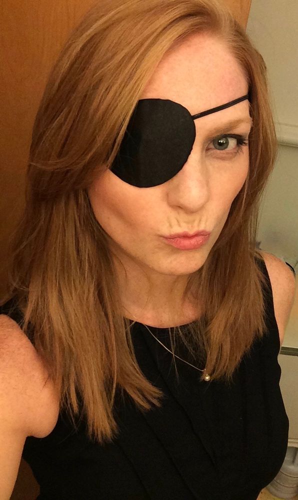 Liz wears a black eyepatch and black costume in this selfie. She has shoulder length y hair.