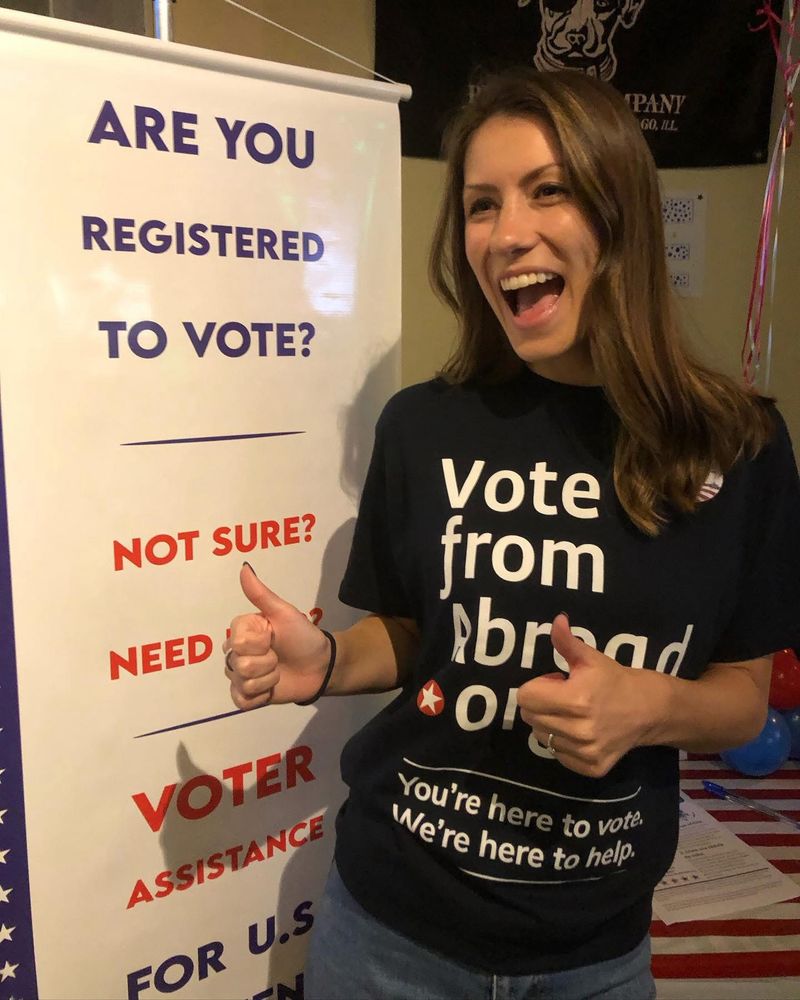 A woman wears a VoteFromAbroad.org T-shirt in front of a VoteFromAbroad.org sign and gives two thumbs up