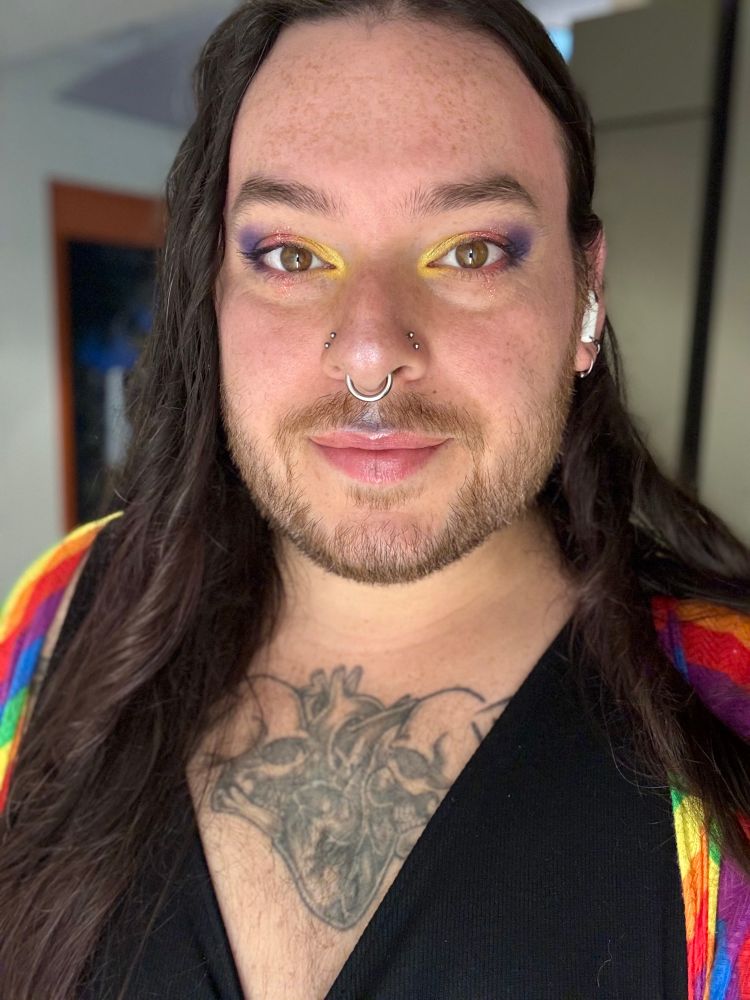 Person wearing a black v neck dress and rainbow duster with long brown hair and colorful makeup in a selfie. 