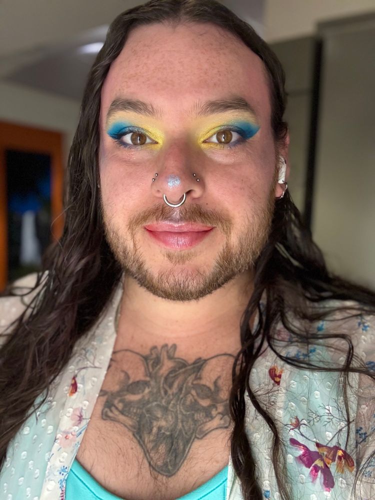 Person with colorful makeup and long brown hair wearing a teal jumpsuit and a flowery shawl over it in a selfie. 