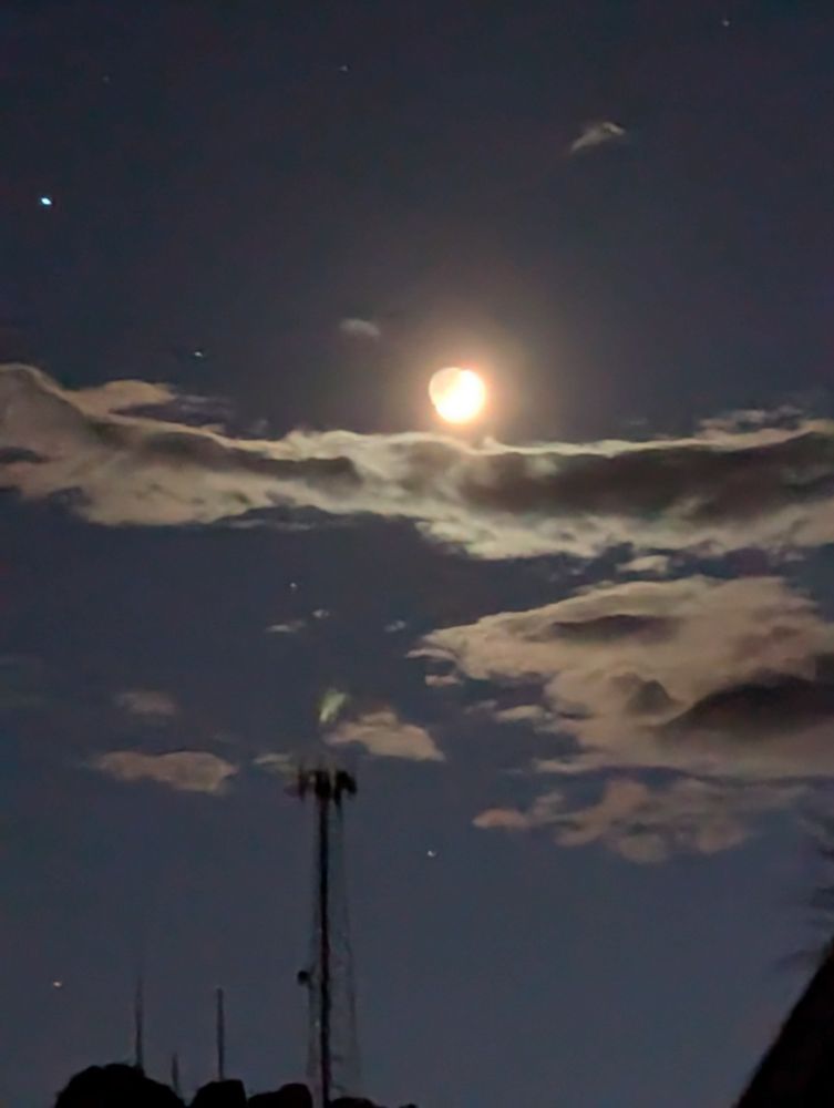 Original Astro-Photography by G of Real Nature Magic. The crescent moon with Earthshine effect rises over a few wispy clouds in the night sky. Regulus is a bright point to the left, rising over the clouds. A radio tower is silhouetted beneath.