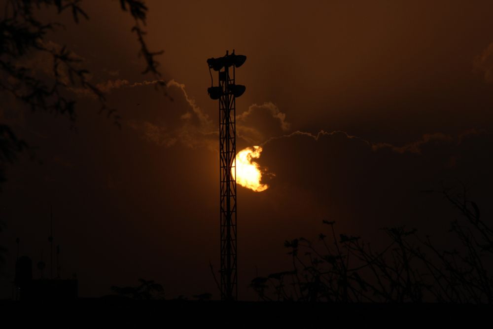 Photo of the sunset seen really close. The image is dark and in the middle there's a darkened tower next to the Sun in the middle of the picture. Some clouds are covering the sun partially. In the upper left and lower right corners there are some darkened trees
