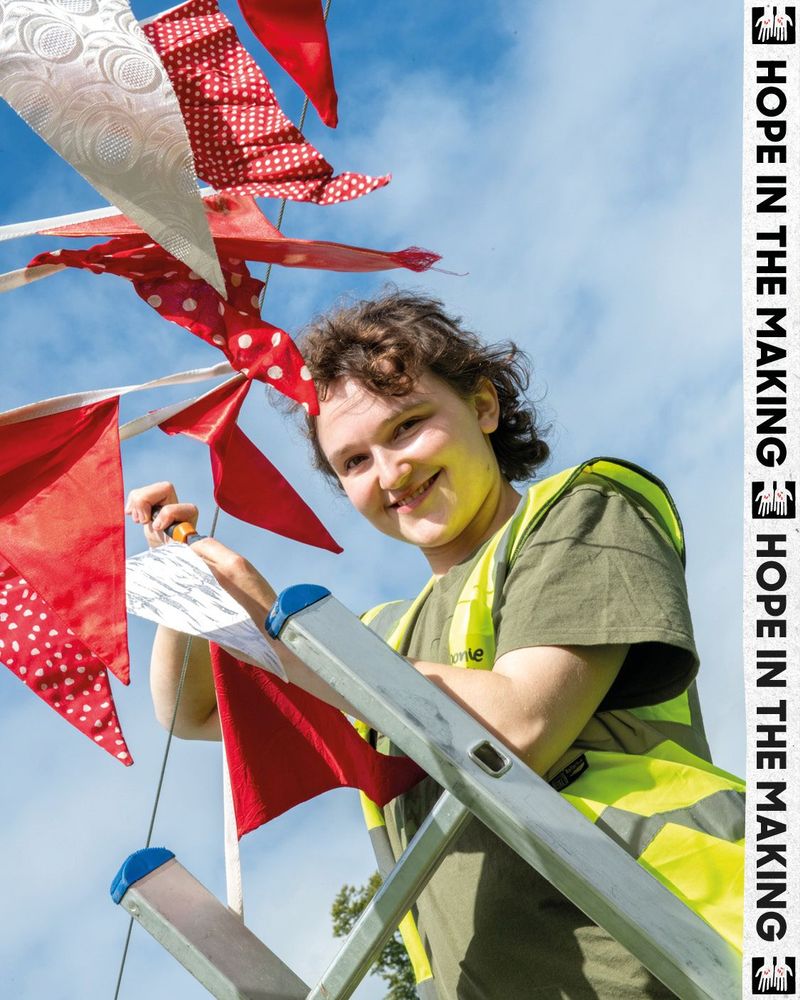Smiling volunteer in yellow hi-vis vest stands on a ladder, hanging red and white bunting against a bright blue sky.