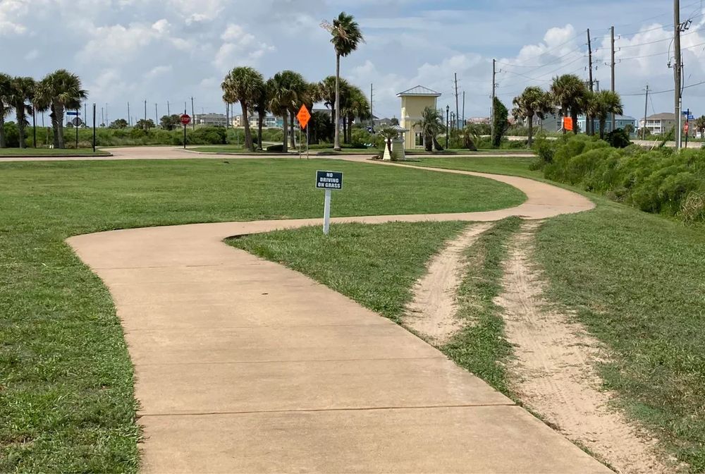 A path worn away in grass that goes in a straight line towards the other side of the park, bypassing the winding paved path