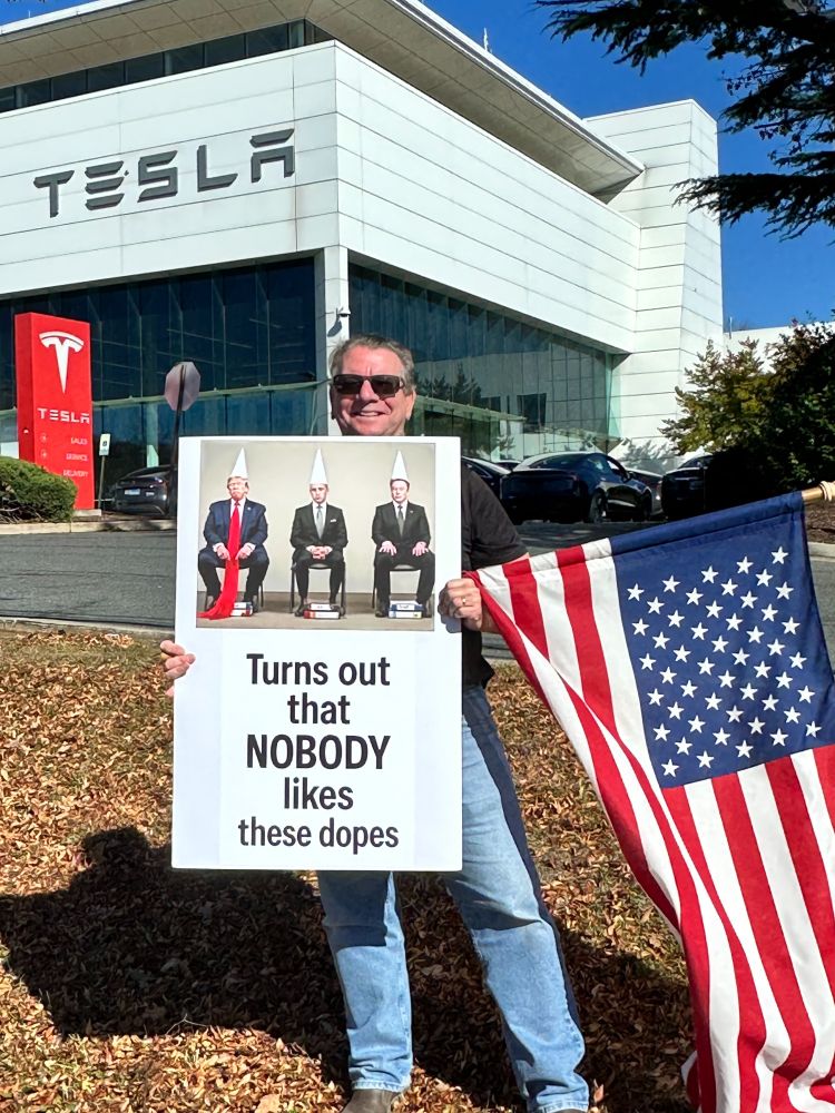 Photo of many holding a sign and American flag in front of the Tesla showroom. The sign depicts Trump, Stephen Miller, and Elon Musk seated and wearing white dunce caps. Text reads, turns out that nobody likes these dopes.