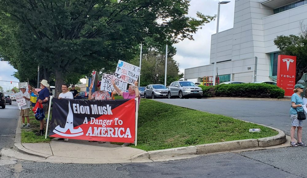 Many people standing on a sidewalk in front of a Tesla showroom. A banner in front reads, Elon Musk, a danger to America.