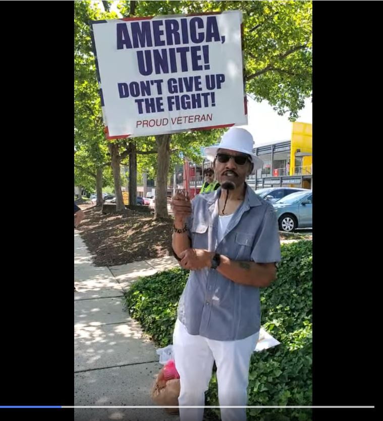 Still frame from a video showing a man, standing on a sidewalk in front of some stores, at a Tesla Takedown rally in Rockville, MD. He is holding a sign that read, "America! Unite! Don't give up the fight! Proud veteran" and wearing dark glasses, white hat, gray shirt, and white pants.