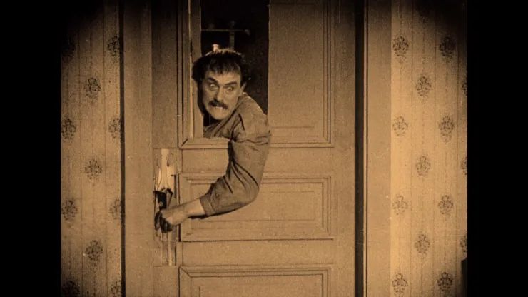Fotograma de la película "La carreta fantasma", en tono sepia. El fotograma muestra a un hombre con bigote y gesto desquiciado intentando cruzar el umbral de una puerta medio rota, asomando desde el otro lado y aferrando el pomo con su mano izquierda.