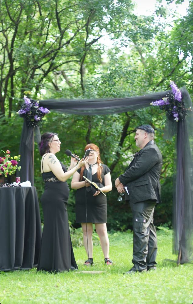 Matty and Stef stand under a black tulle-draped wedding arch adorned with purple and black floral arrangements, exchanging vows during an outdoor ceremony. Stef, wearing a fitted black gown with lace details and purple streaks in her hair, holds a microphone and a small booklet. The groom, dressed in a black patterned jacket and leather pants, smiles warmly while holding his own vow book and sunglasses. Between them stands the officiant in a black dress, holding a notebook and a mic. A small black-draped table with flowers and ceremony items is visible to the side. The scene is set in a lush, wooded area.