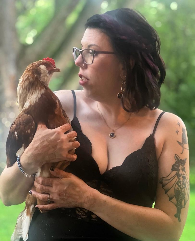 Stef, in a black lace dress, holds a brown and yellow chicken in her arms while making a playful, expressive face directed at the bird. She wears glasses, dangling black stone earrings, and a matching pendant necklace. Her short dark hair has purple streaks, and her arm features a bold tattoo of a retro-style bat-winged woman. The background is softly blurred greenery, suggesting an outdoor setting.