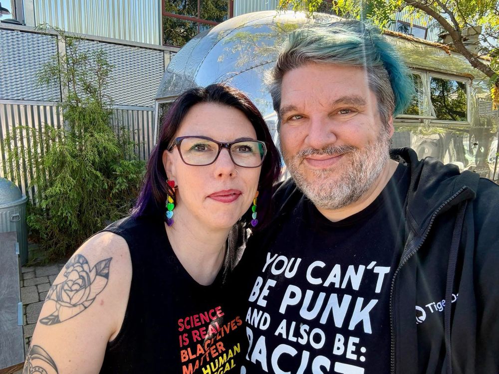  A smiling couple stands outside at a local pride fest. The woman has black hair with  purple highlights, wears glasses, colorful rainbow heart dangling earrings, and a sleeveless black shirt that reads “Science is real, Black Lives Matter, No human is illegal.” She has a floral tattoo on her upper arm. The man beside her has short teal-tinted hair, a trimmed beard, and a black hoodie over a T-shirt that says “You can’t be punk and also be racist.” Behind them are trees, an Airstream trailer, and a corrugated-metal building gleaming in the sunlight.