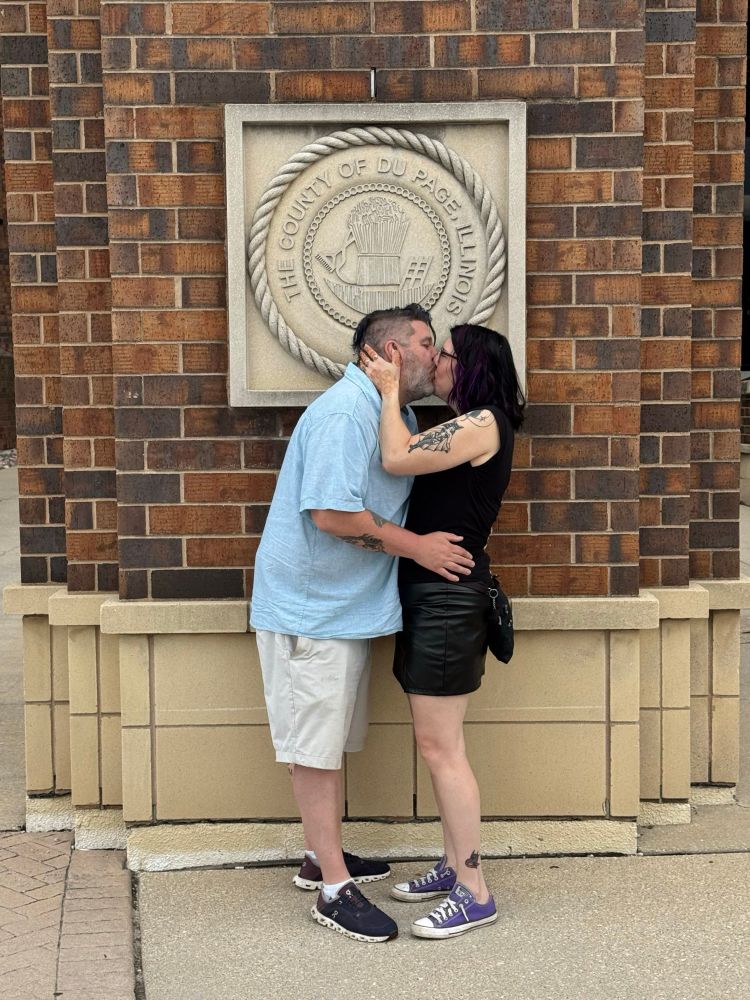 A couple shares a kiss in front of a brick wall with a stone seal that reads “The County of DuPage Illinois.” The man wears a light blue button-up shirt, tan shorts, and navy sneakers. The woman wears a black top and black leather miniskirt with purple Converse sneakers. She has visible tattoos on her arm and ankle, and her dark hair is streaked with purple. They are standing closely, embracing affectionately. 