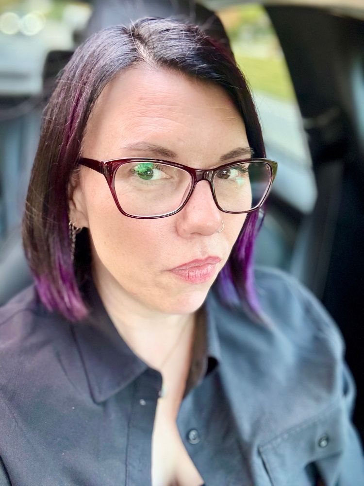 A woman with chin-length black hair that has purple highlights, wearing maroon glasses and a black button-up shirt, sits in a car and looks toward the camera with a neutral expression. The background is softly blurred, showing muted outdoor colors through the car window.