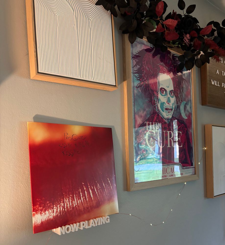 A wall display featuring a framed vinyl record of The Cure’s Kiss Me, Kiss Me, Kiss Me album on a “Now Playing” stand, next to a vibrant, framed concert poster for The Cure’s 2023 show at the United Center in Chicago, illustrated in a gothic, skeletal style. Above the poster is a garland of black and deep red faux flowers. The arrangement is surrounded by minimalist textured artwork in natural wood frames, and a string of soft fairy lights runs across the wall.