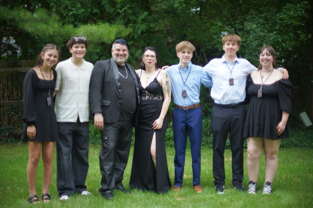 A blended family stands together on a grassy lawn in front of a backdrop of trees, dressed in semi-formal black and white attire for a special occasion. The couple at the center wear coordinated black outfits—the woman in a fitted gown with a lace bodice and slit, and the man in a textured jacket, satin lapels, and black leather pants. Surrounding them are five teenagers, all smiling and wearing laminated backstage pass-style lanyards, adding a playful, music-themed touch to the event. The setting and attire suggest a rock-inspired wedding celebration.