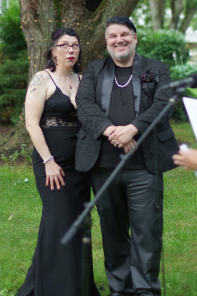Matty and Stef stand outdoors in front of a large tree wrapped with string lights. Stef wears a fitted black gown with a lace bodice, glasses, and bold red lipstick. She is trying not to cry while listening to her sister speak. Matty is smiling warmly, dressed in a textured black jacket, black shirt, and leather pants, accessorized with a chain necklace and boutonnière. A microphone and someone’s hand holding papers are visible in the foreground.