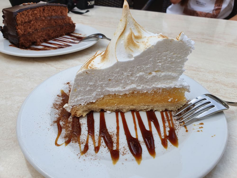 Auf einem weißen, mit flüssigen  Schokoladenstreifen verzierten Teller befindet sich ein Stück Kuchen: dünner Boden, gelbe Frucht Masse, darüber eine dicke Schicht weißes Baiser, das in eine Spitze nach oben geformt wurde und leicht gebräunt wurde.
Eine Gabel wurde auf den Teller gelegt. In Hintergrund ein Teller mit Schokoladenkuchen. 