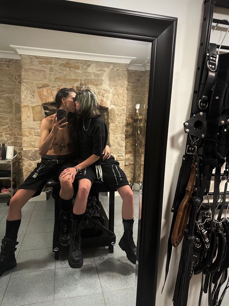 Taken from the same mirror photo angle in the sandstone dungeon, Nyx now takes the mirror photo as she and Cleo sit widespread atop the same padded table, arms looped around each other and engaged in an eager kiss. No longer in lingerie but still in boots, Cleo wears black dyke shorts and an oversized black tshirt, whilst Nyx is topless, also wearing black dyke shirts with a carabiner full of keys laying down her leg.

