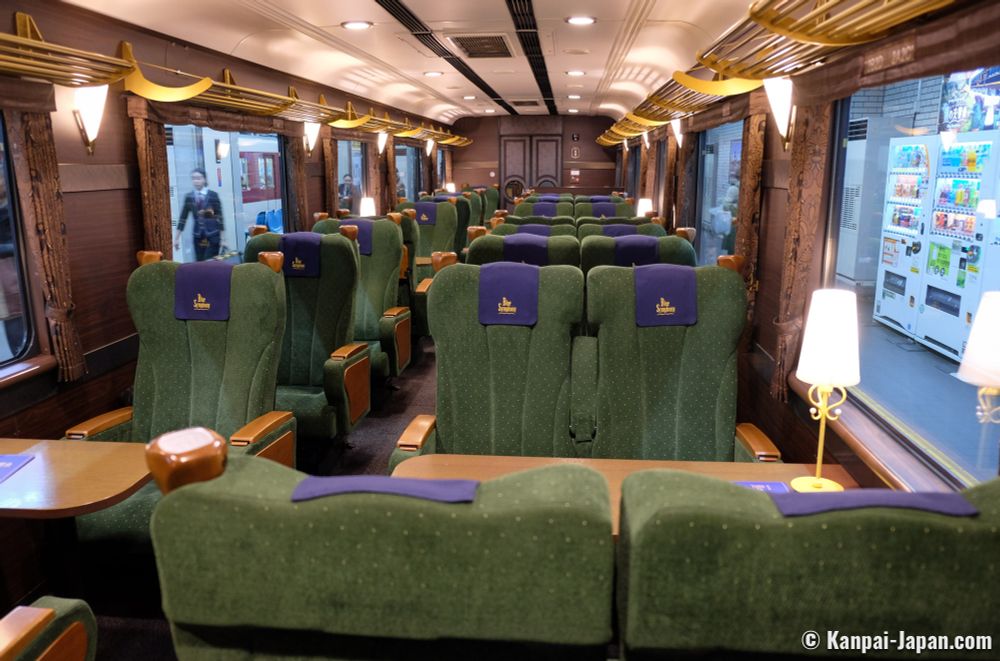Interior of the Blue Symphony, a limited express train operated by Kintetsu connecting Osaka-Abenobashi and Yoshino station in Nara prefecture. Composed of only three cars, this touristic train offers its passengers the experience of a comfortable and peaceful travel throughout the Japanese countryside. The seats are fern green with purple antimacassars. The table between the facing seats has a brass lamp with a narrow white shade. 