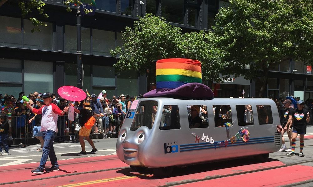 A man carrying a pink parasol walking in front of an anthropomorphic BART car wearing a giant pride rainbow top hat at the San Francisco Pride Parade.

Very Tingleverse. 
