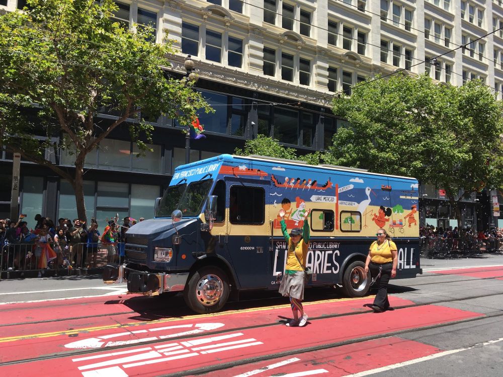 A bookmobile painted with a beach scene and text reading Libraries for All accompanied by two parade walkers wearing yellow shirts