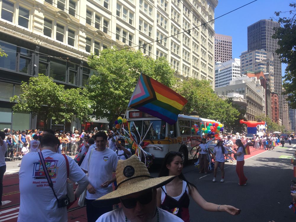A vintage MUNI bus surrounded by parade walkers balloons and a large pride foreward flag