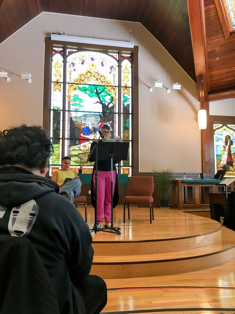 Annalee Newitz in a dark blue tailcoat, and magenta tuxedo trousers with a contrast stripe, and silver shoes, reads from a lectern on a highly polished wood floor two step dais in a church in front of a stained glass window depicting the story of the good samaritan. Alex Hanna wearing a yellow top and jeans is sitting on a chair to Annalee's right reading along. A person the foreground is wearing a black hoodie  with no face from Spirited Away, so this is obviously a very cool, smart, and fun crowd who likes good things.