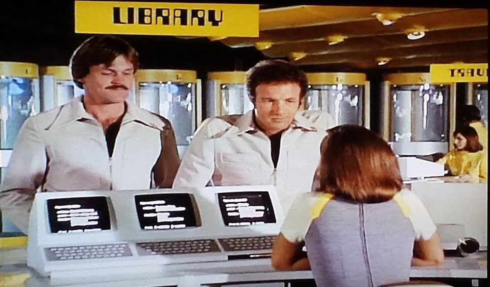 The library Circulation desk in a scene from ‘Rollerball (1975)
Two men in snazzy jumpsuits talk to a woman in front of a series of computer terminals under a yellow and black sign reading LIBRARY in a banging 1970s "computer" typeface