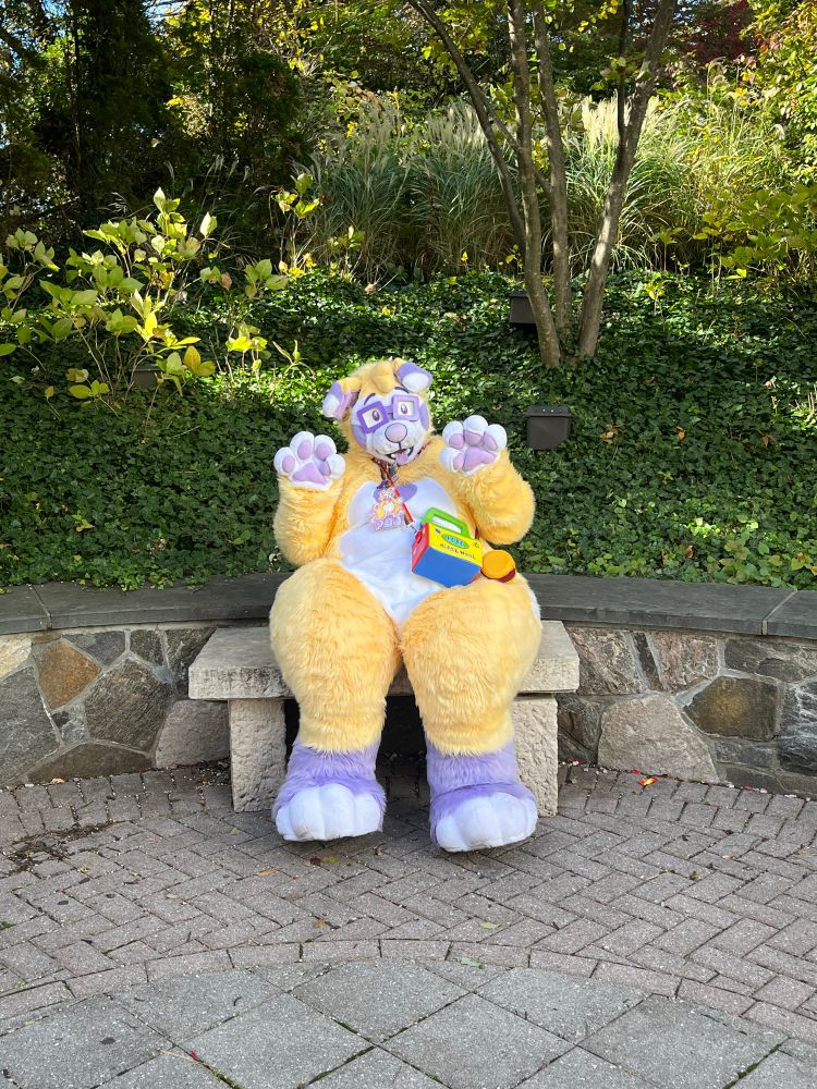 A yellow dog fursuiter sitting on a bench outdoors. 