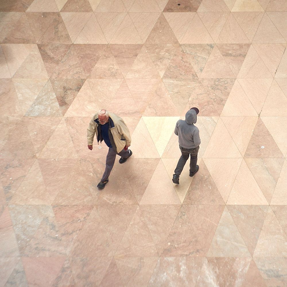 Two people walking side by side in opposite directions. They seem to be following the grout lines of the geometric granite floor tiles. Shot with a Jupiter 8 50mm f2 probably around f5.6 or f8 from the catwalk at The National Gallery East Building 