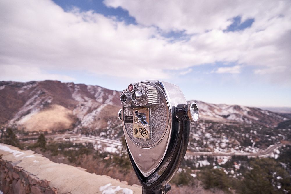 A closeup image of a Tower Optical coin operated binocular viewer overlooking some snow covered mountains under a partly cloudy sky. 
