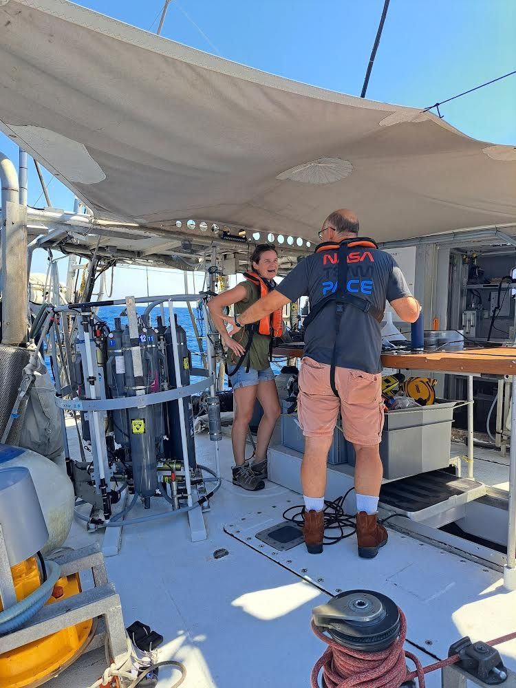 two scientists on a research vessel talking, standing next to a CTD rosette. 