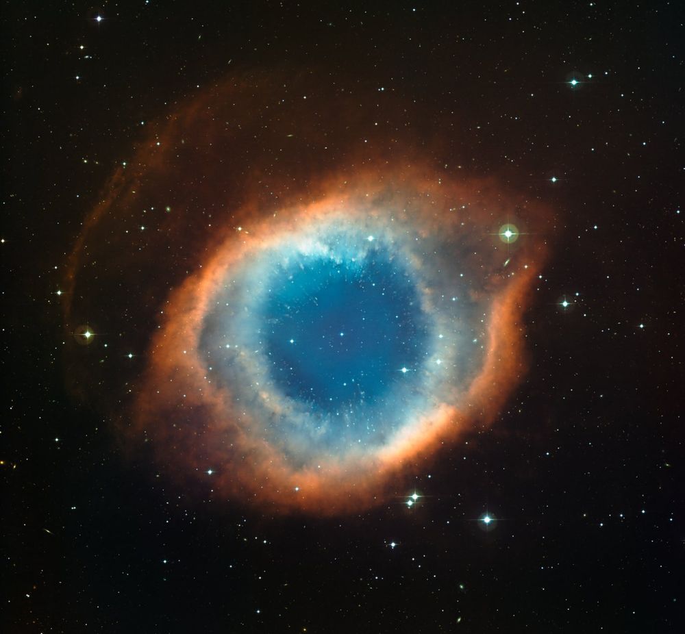A vivid image of a nebula, showing a bright blue centre surrounded by a glowing ring of reddish-orange gas and dust, set against a backdrop of stars in deep space.