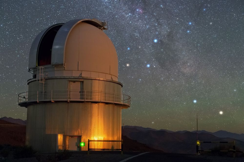 A close-up view of a large observatory telescope dome under a brilliant night sky filled with stars. The dome is partially open, with soft orange lighting illuminating the exterior. Distant mountains are silhouetted against the horizon.