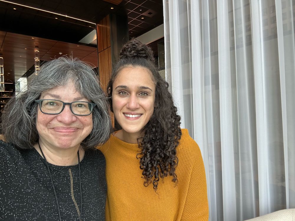 Two smiling women one in graying chin length hair and glasses and the other with long curly dark hair, standing next to a white curtain 