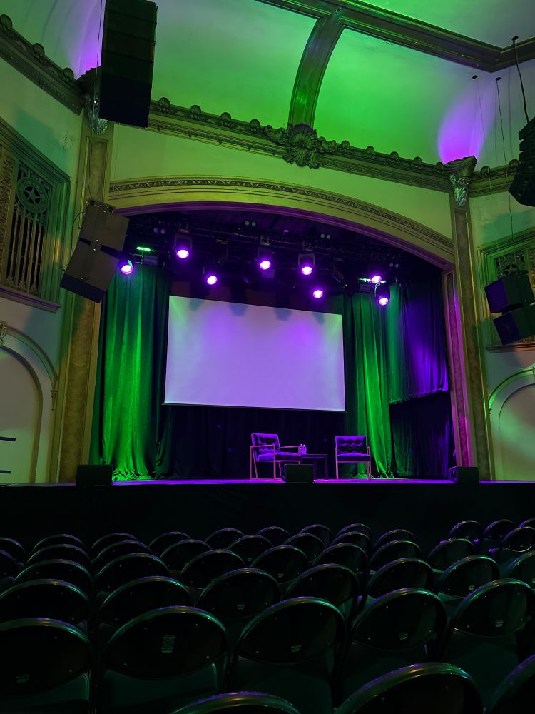 A picture of a movie theater, the lights and green and purple. Two chairs are on a stage in front of the screen. 