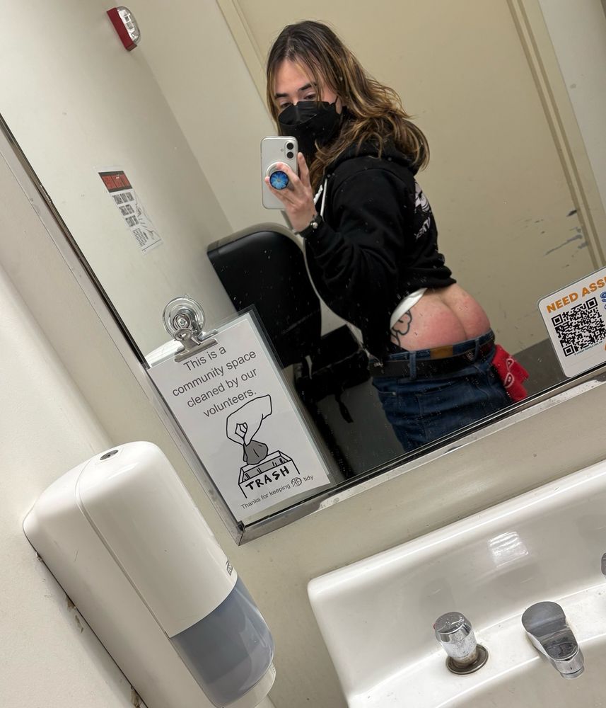 A photo of Dusty reflected in a bathroom mirror, looking over his shoulder. His jeans are pulled down, showing off his red ass from a recent beating. He’s wearing a black mask, black hoodie, and red hanky in the left pocket.