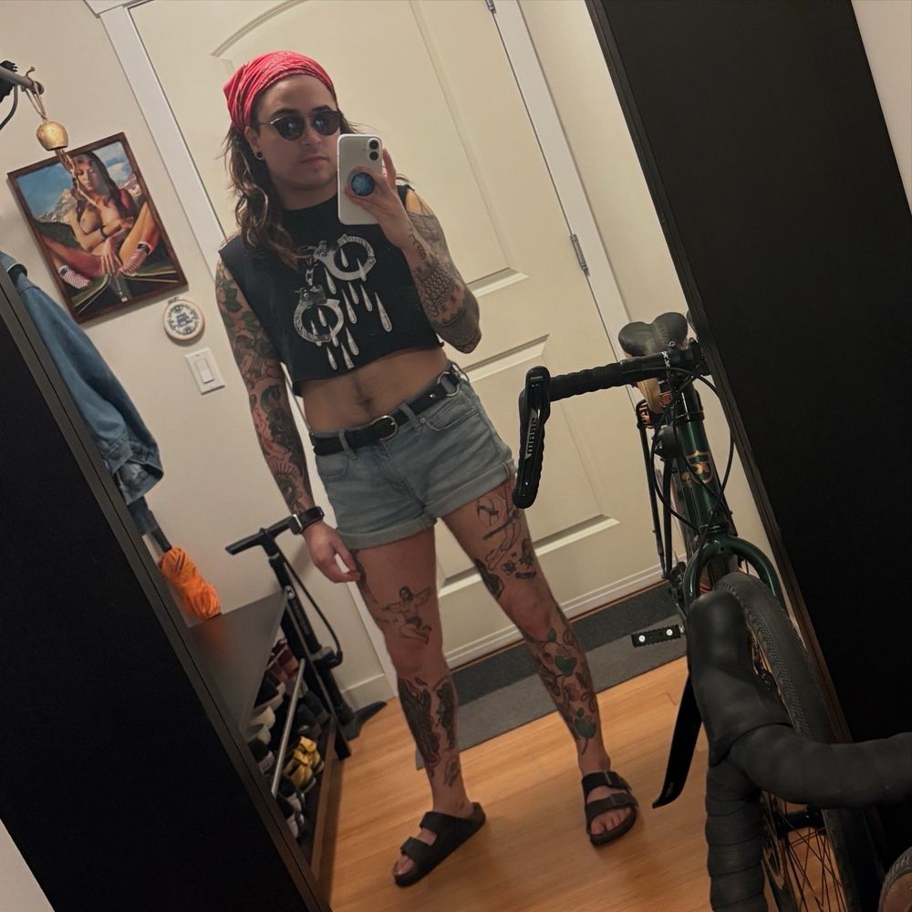 A mirror selfie of Dusty’s outfit: a red bandana tying his hair back, sunglasses, a black sleeveless crop top that’s printed with a pair of handcuffs dripping blood, light wash denim shorts and a black leather belt with ring details, and black leather Birkenstocks.