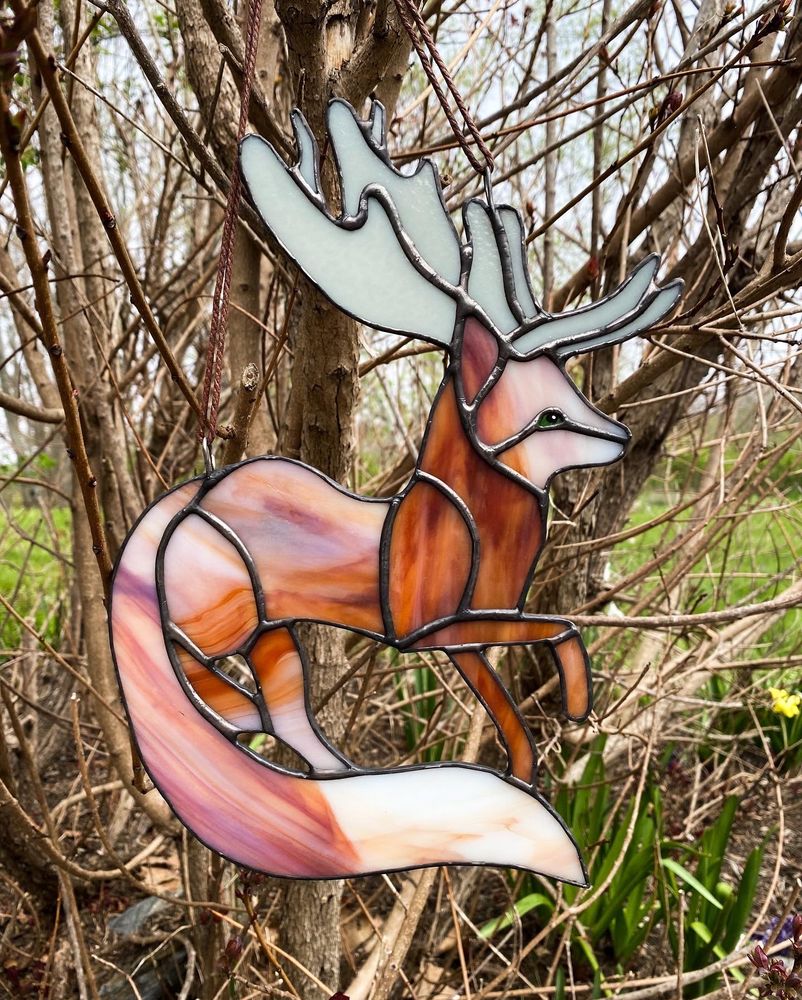 Stained glass fox with luxurious tale and a rad of antlers. It hangs from a small tree branch outdoors 