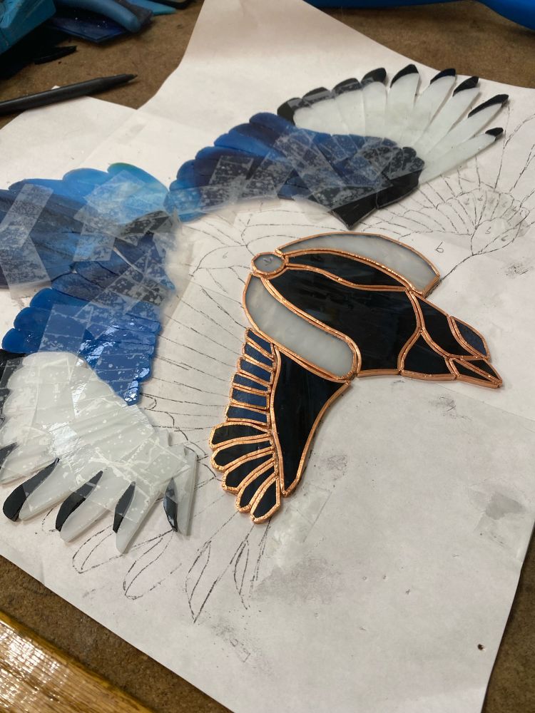 WIP stained glass magpie. Its wings are spread and it’s made of 108 hand cut pieces of glass. The body and part of one wing are copper foiled while the rest is held together with bits of scotch tape. 