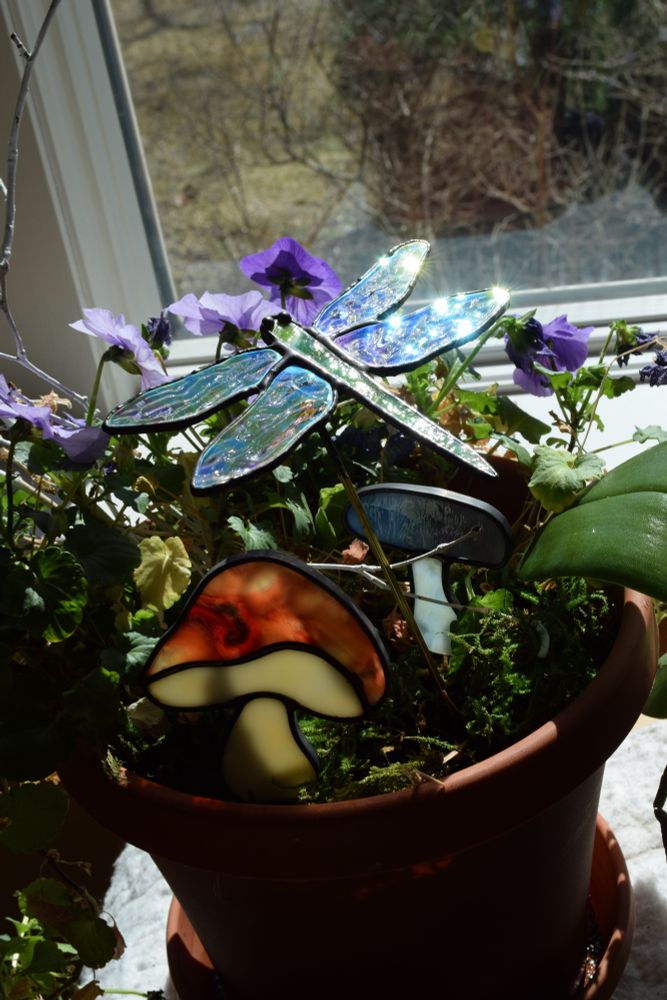 Flowerpots in a window with two stained glass mushrooms and a dragonfly on little garden stakes in the pots among leaves and flowers. The dragonfly has iridescent wings and they sparkle in the sunlight.  