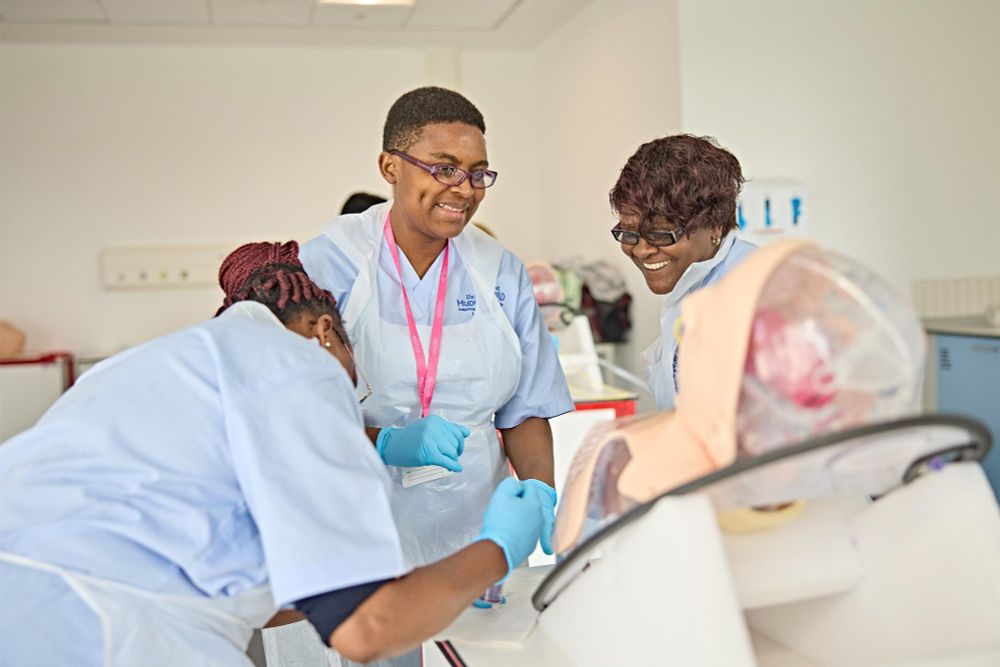 Three healthcare professionals are interacting and smiling around a medical training mannequin in a clinical setting.