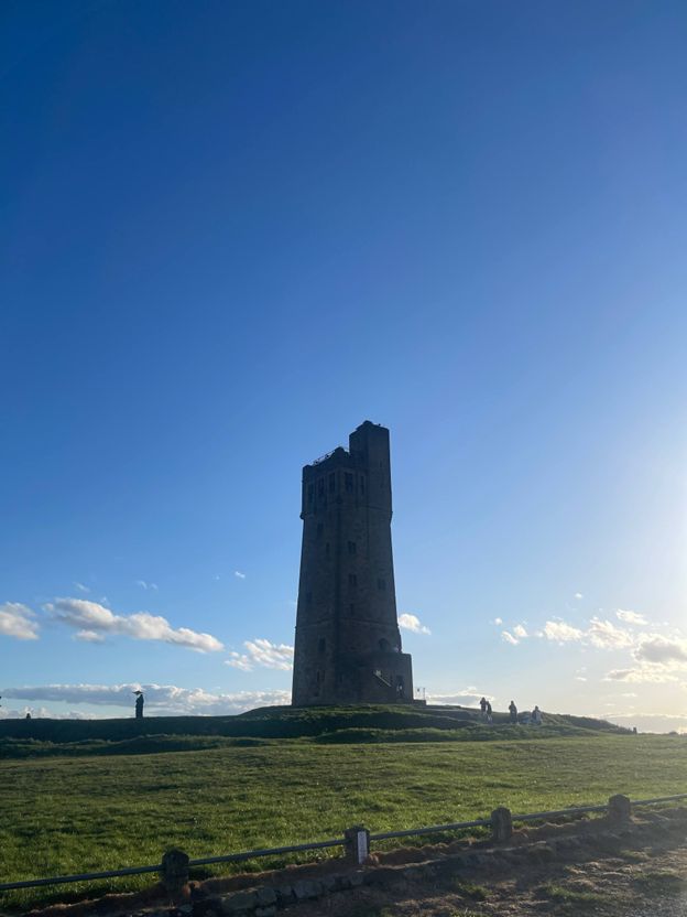 Victoria Tower on Castle Hill stands under a bright blue sky with sunbeams, surrounded by open grassy fields and a few visitors in the distance.